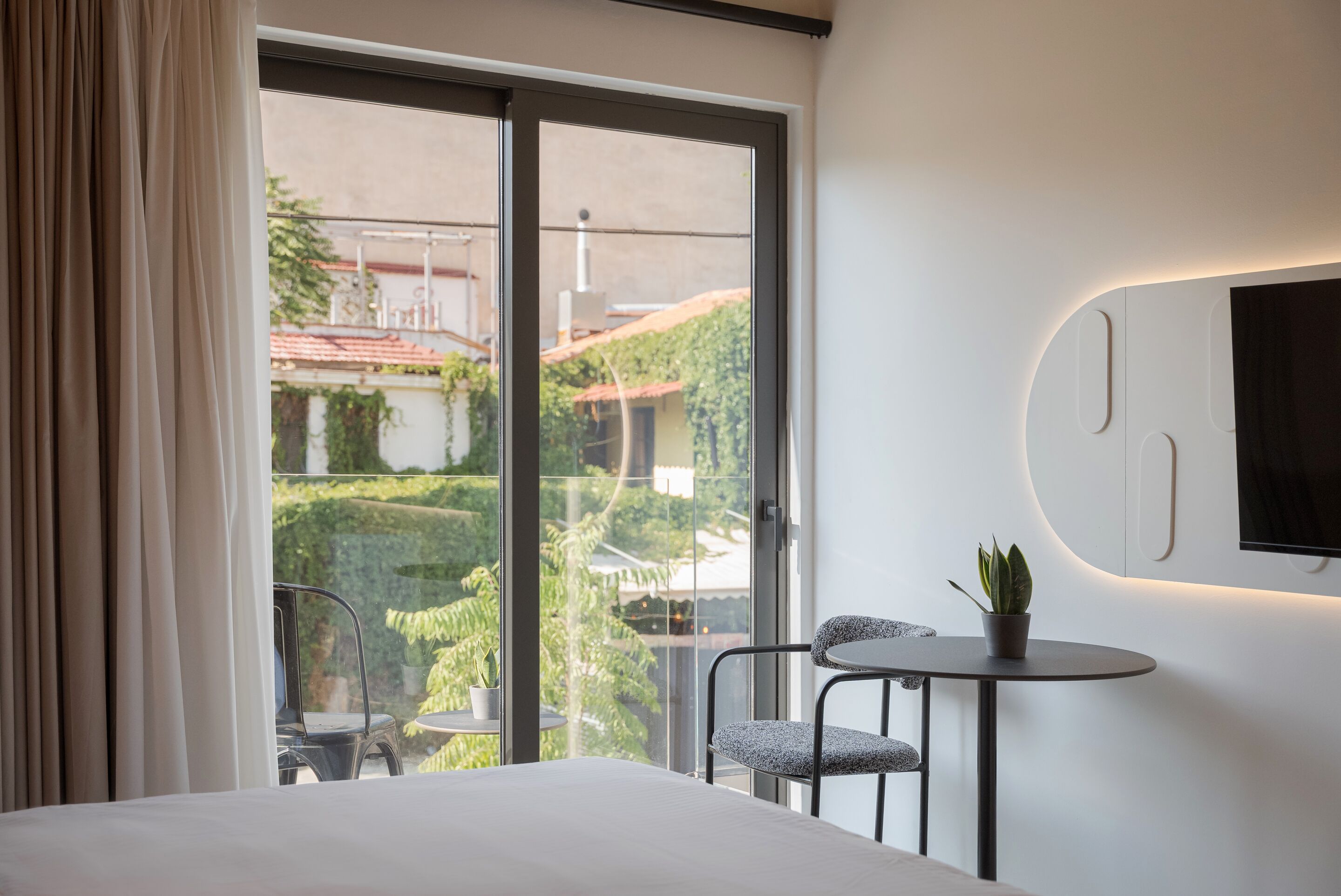 Et moderne hotelværelse med en stor skydedør til en balkon, et rundt bord med en stol, en potteplante og et ophængt fjernsyn på væggen.
