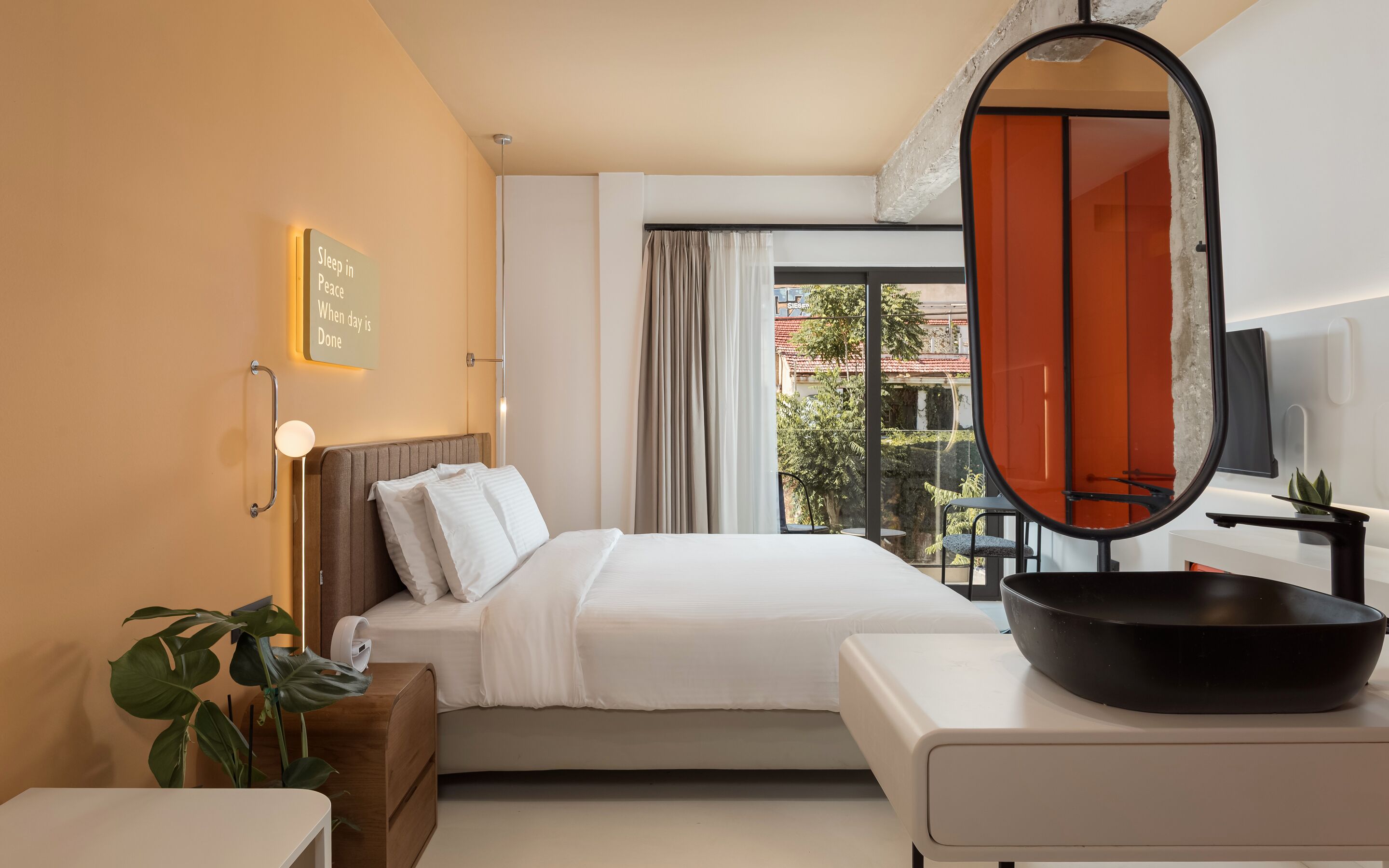A hotel room with a bed, bedside table, a mirror, a window with curtains, and a view of the outside.