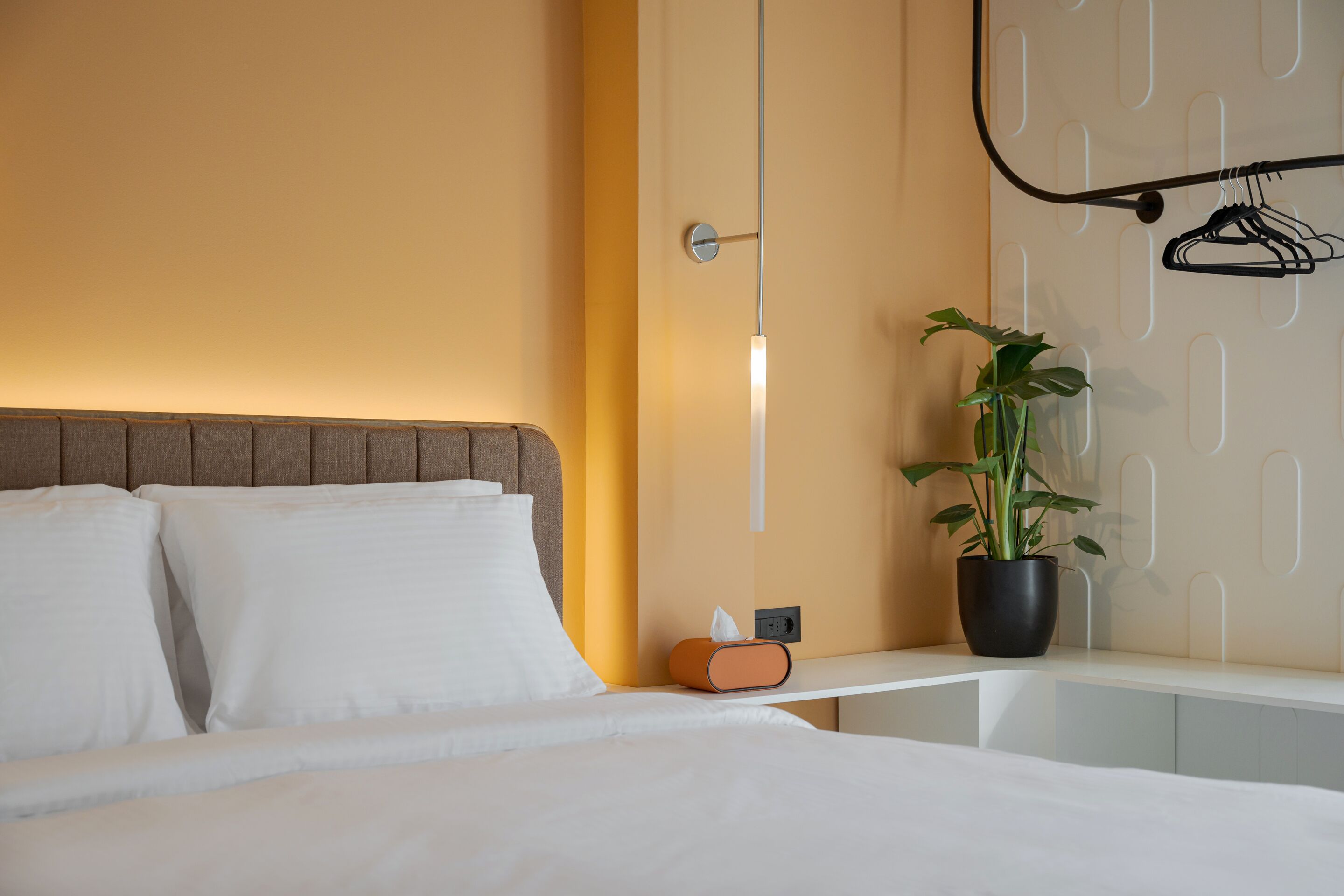 A hotel room with a bed, a bedside table, a plant, and a coat rack.