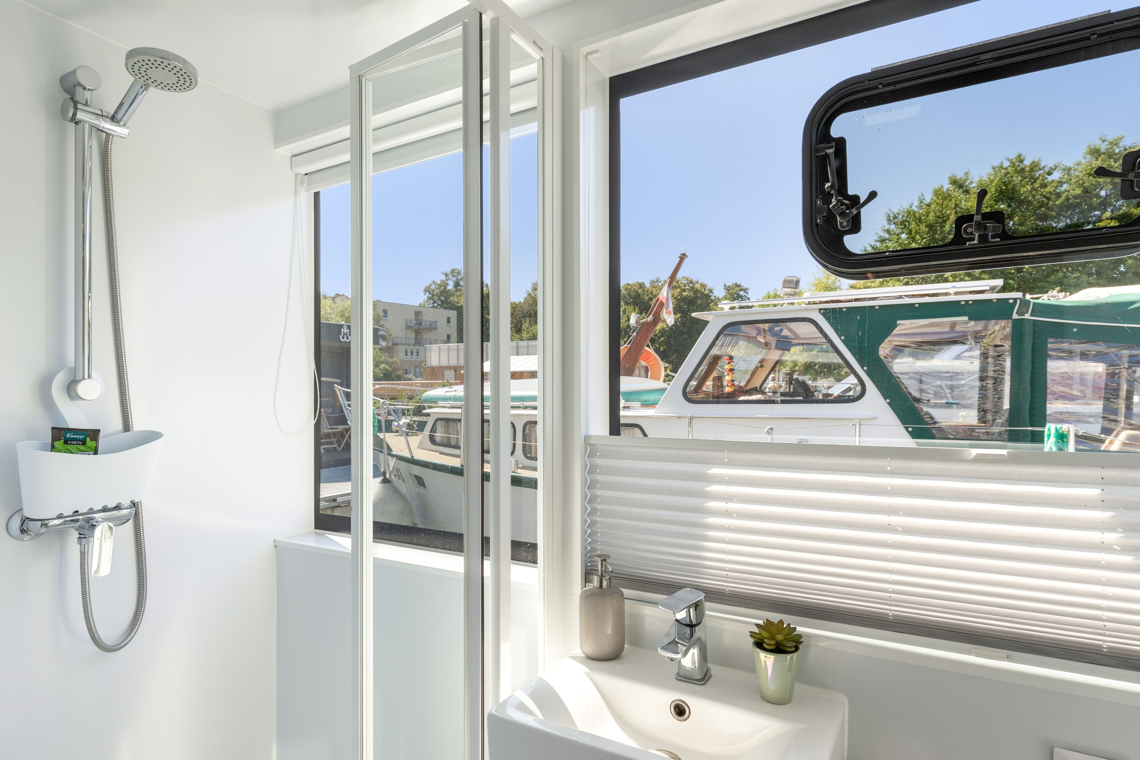 A bathroom with a shower, a sink, and a window with a view of boats and buildings.