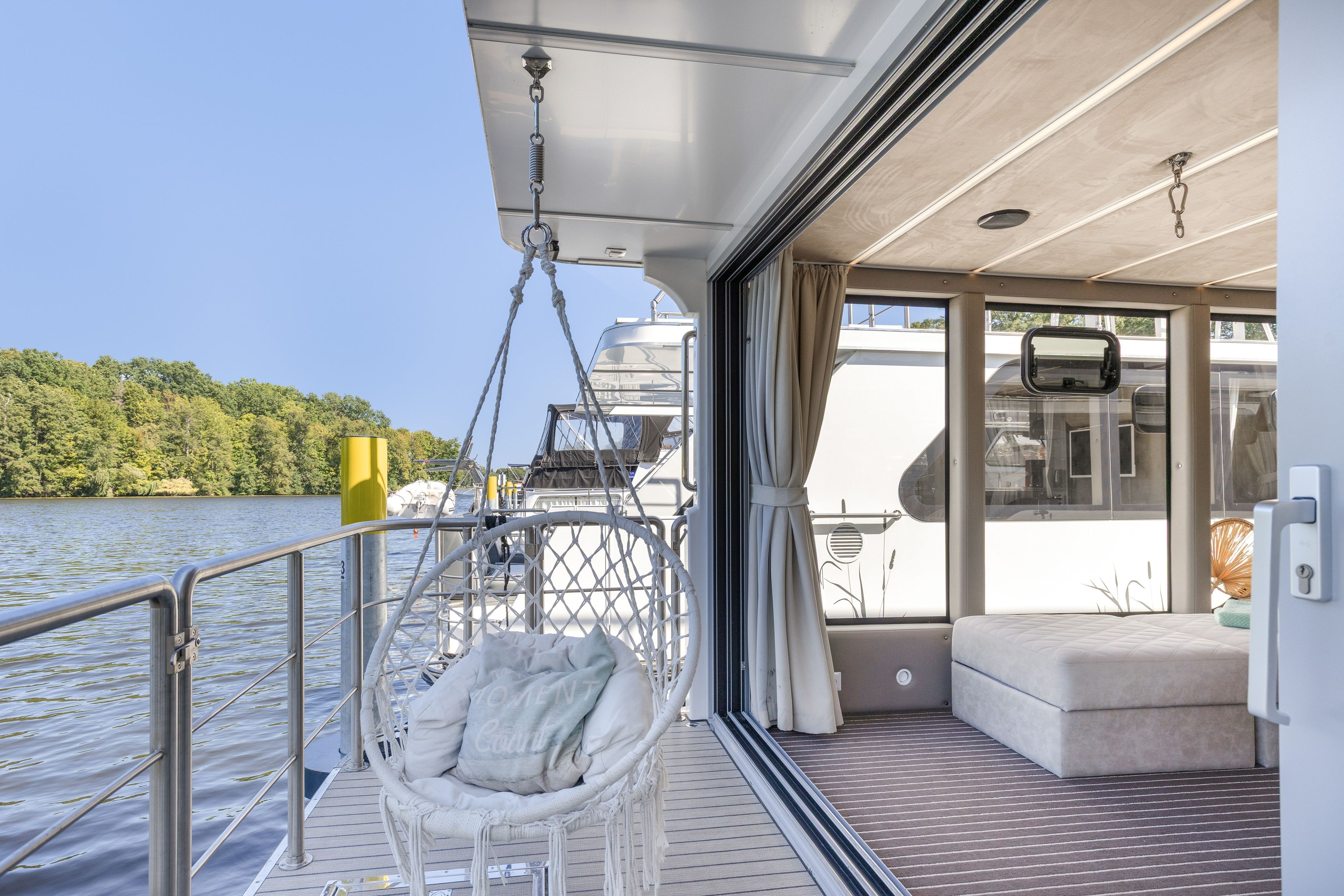 Een scheepsdek met een hangstoel, een comfortabele bank en uitzicht op water en bomen.