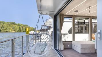 Een scheepsdek met een hangstoel, een comfortabele bank en uitzicht op water en bomen.