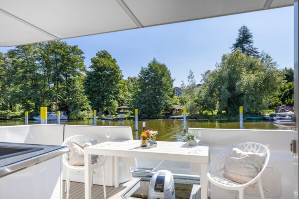 Cubierta de barco con una mesa preparada para dos, rodeada de sillas de mimbre blanco, con vista a un río tranquilo con árboles y edificios al fondo.