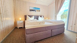 A bedroom with a large bed, wooden walls, a nightstand with a lamp, and a window with a view. - Holiday Home 'Wohlfühlhaus an der Müritz' with Wi-Fi (Rechlin)
