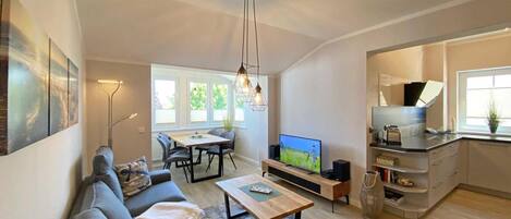 A modern living room with a sofa, coffee table, and TV. There is a dining area with chairs and a table. The kitchen is visible with cabinets and a window.