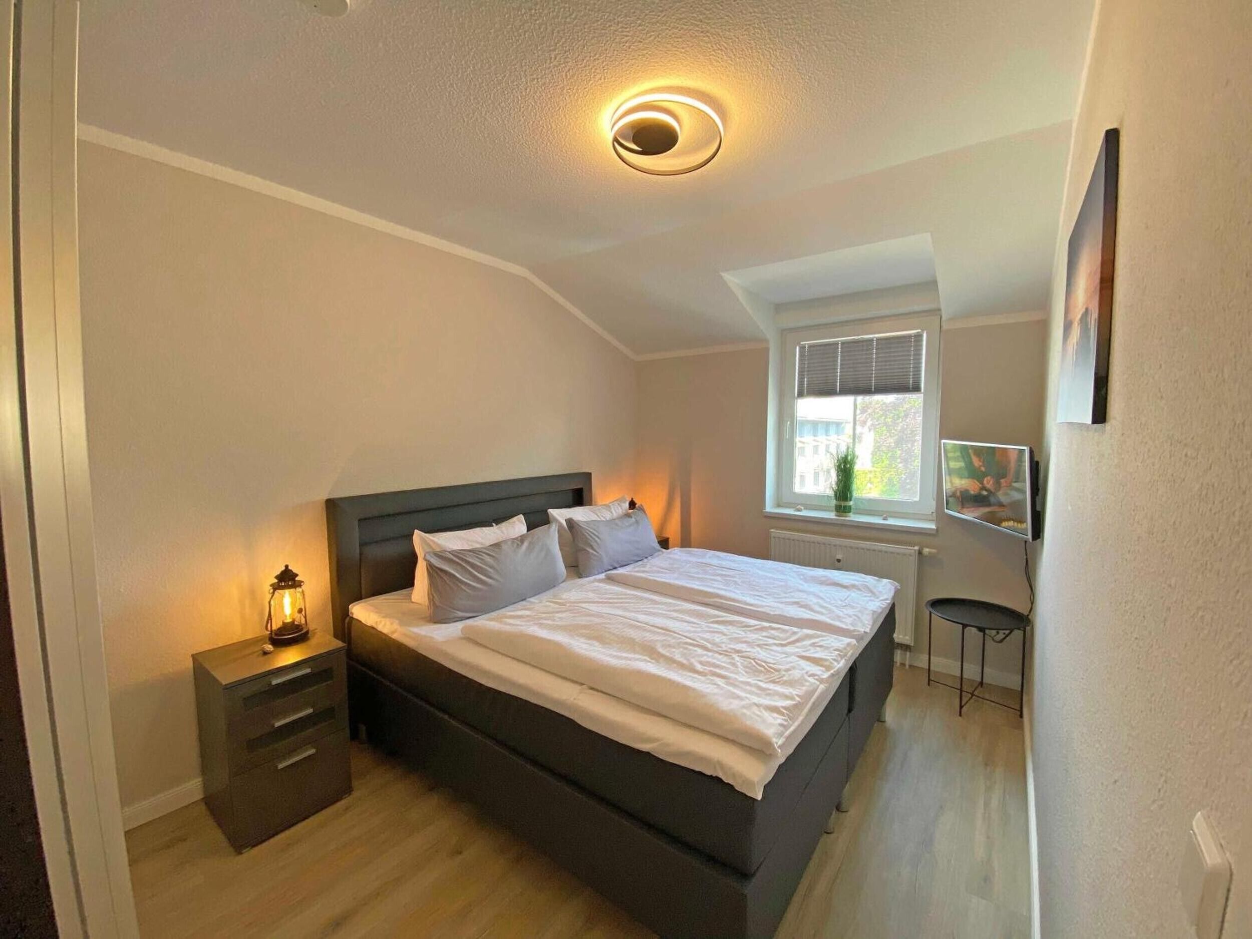 Ein Schlafzimmer mit Bett, Nachttisch, Lampe, Fenster und einem an der Wand befestigten Fernseher.