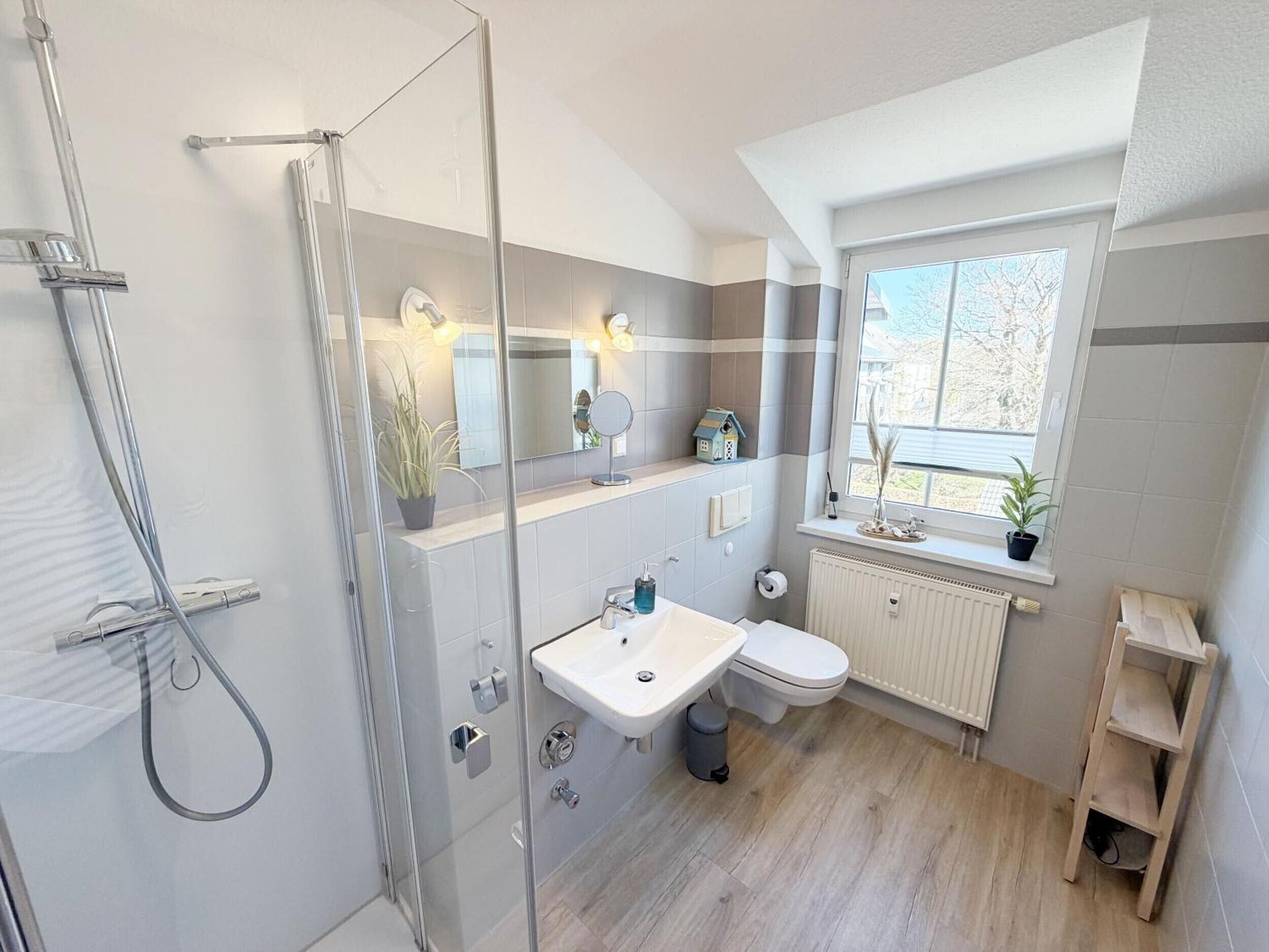 Ein modernes Badezimmer mit einer Duschkabine aus Glas, einem weißen Waschbecken und einer Toilette. Ein Fenster sorgt für natürliches Licht und einen Ausblick ins Freie.