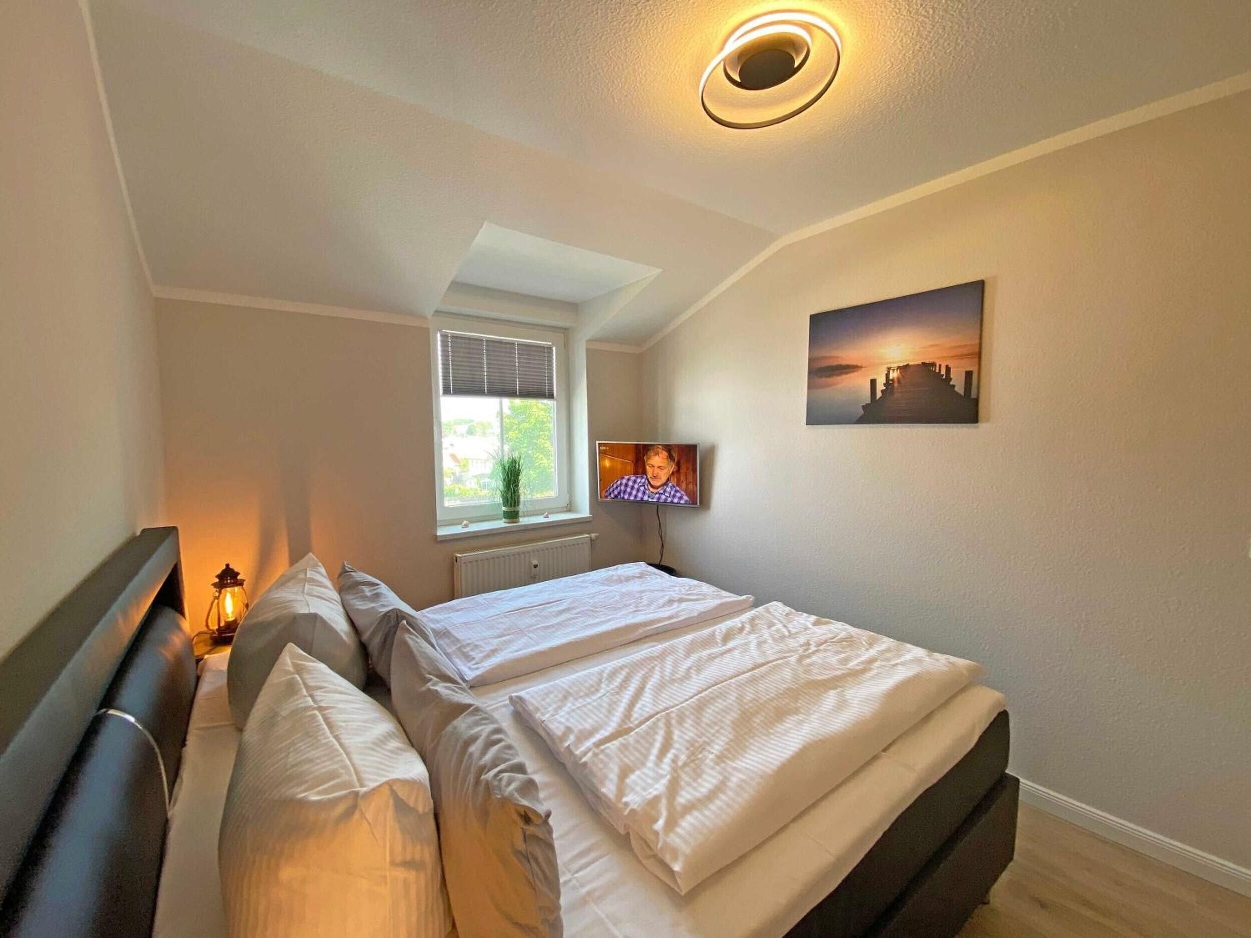 Ein Schlafzimmer mit einem Bett, einem Fenster mit Jalousien, einem an der Wand montierten Fernseher und einer Deckenleuchte.
