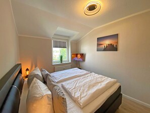 Ein Schlafzimmer mit einem Bett, einem Fenster mit Jalousien, einem an der Wand montierten Fernseher und einer Deckenleuchte.