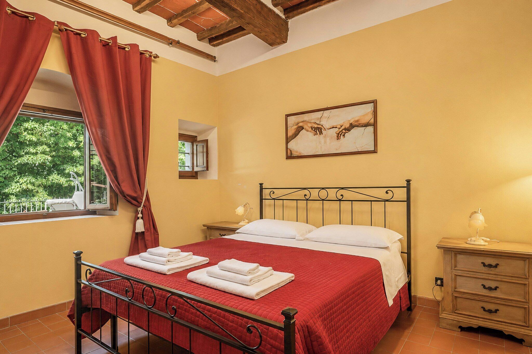 A bedroom with a bed, a nightstand, a window with curtains, a painting, and a view of greenery outside.