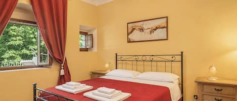 A bedroom with a bed, a nightstand, a window with curtains, a painting, and a view of greenery outside.