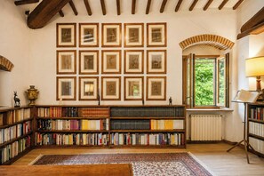 Una biblioteca con estanterías, cuadros en la pared, un radiador y una ventana con vistas a zonas verdes.