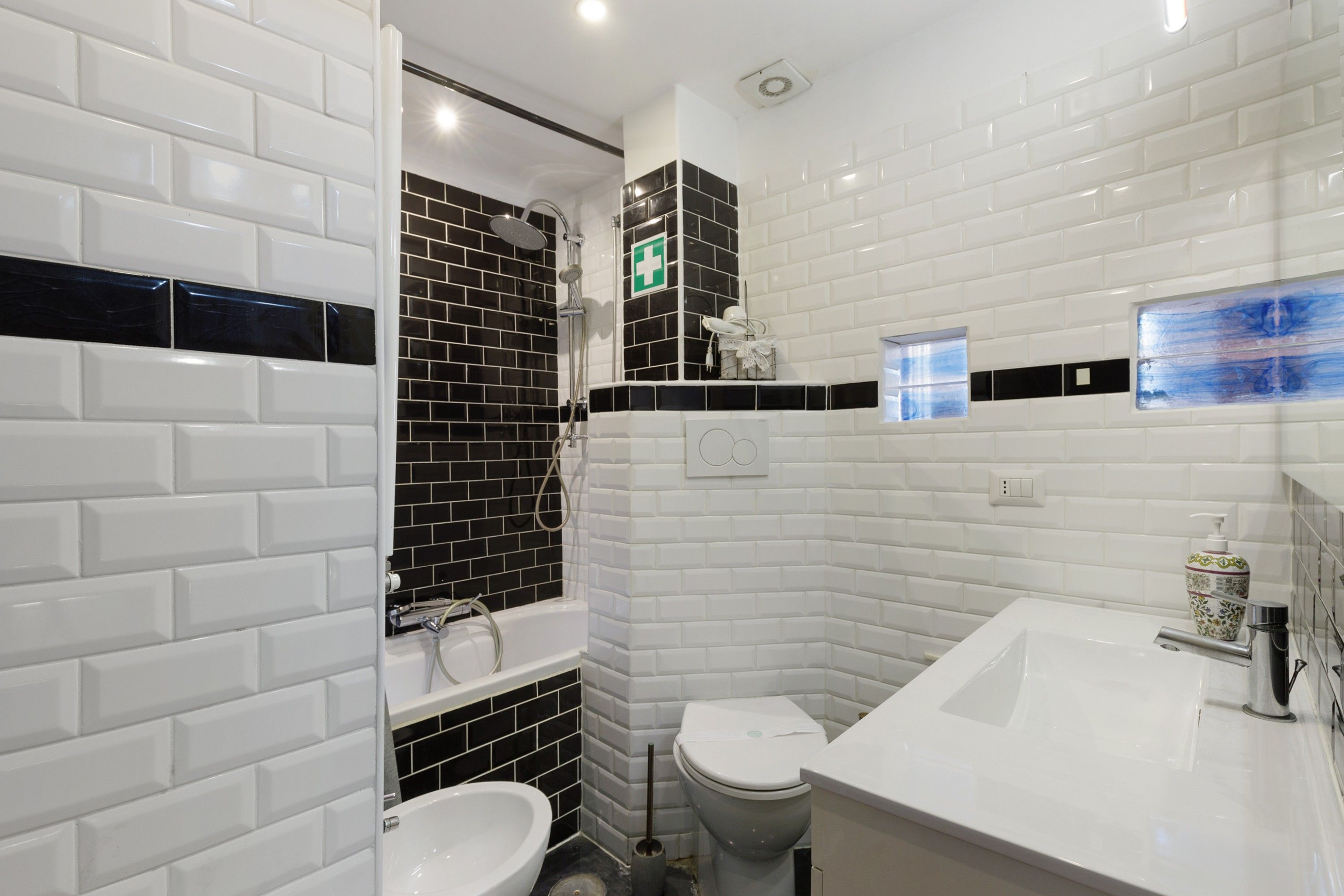 Un bagno con piastrelle bianche tipo metro, un angolo doccia con piastrelle bianche e nere, un water bianco e un lavandino bianco con uno specchio.