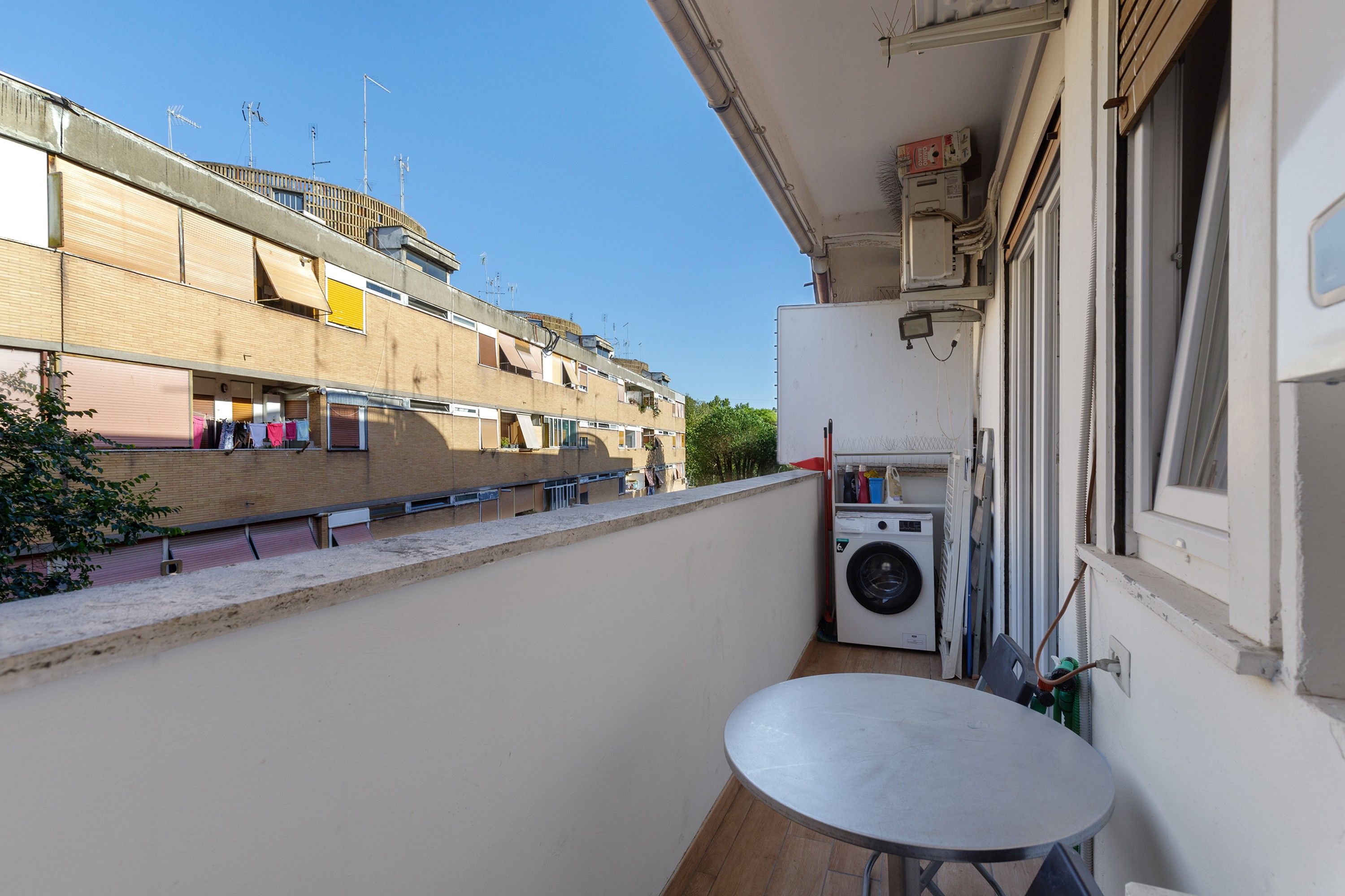 Un balcone con lavatrice, un tavolino e vista sui palazzi vicini.