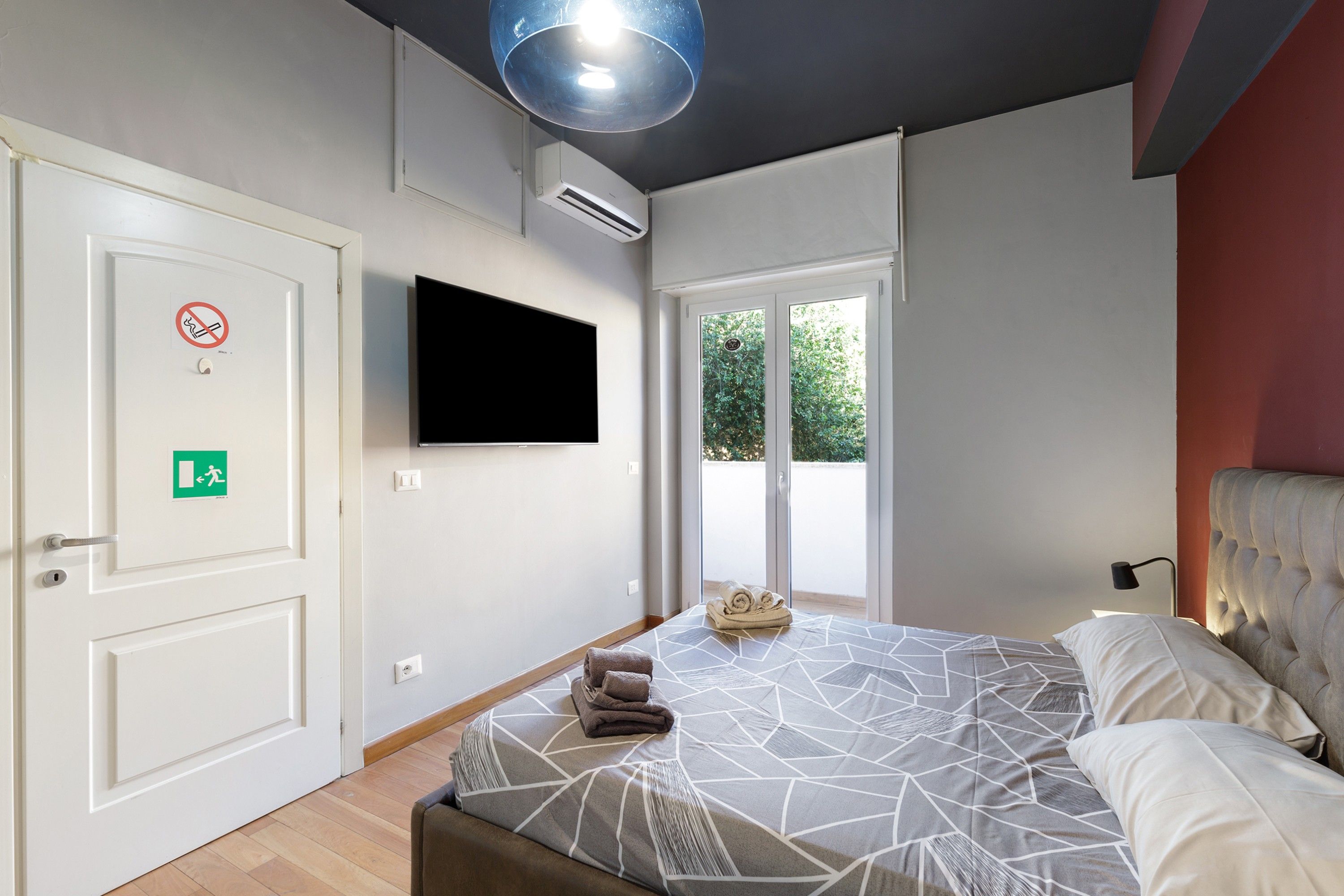 Una camera d'albergo con un letto, una televisione a schermo piatto fissata al muro, una porta con il cartello ‘vietato fumare’ e una finestra che si affaccia sul verde.