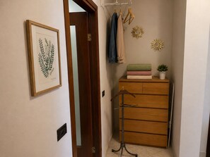 Een gang met een houten deur, een kapstok met ophangers, een radiator en een ingelijst botanisch kunstwerk aan de muur.