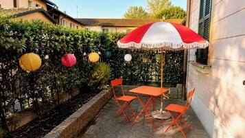 Een terras buiten met een tafel en stoelen, omringd door groen en een decoratieve parasol.