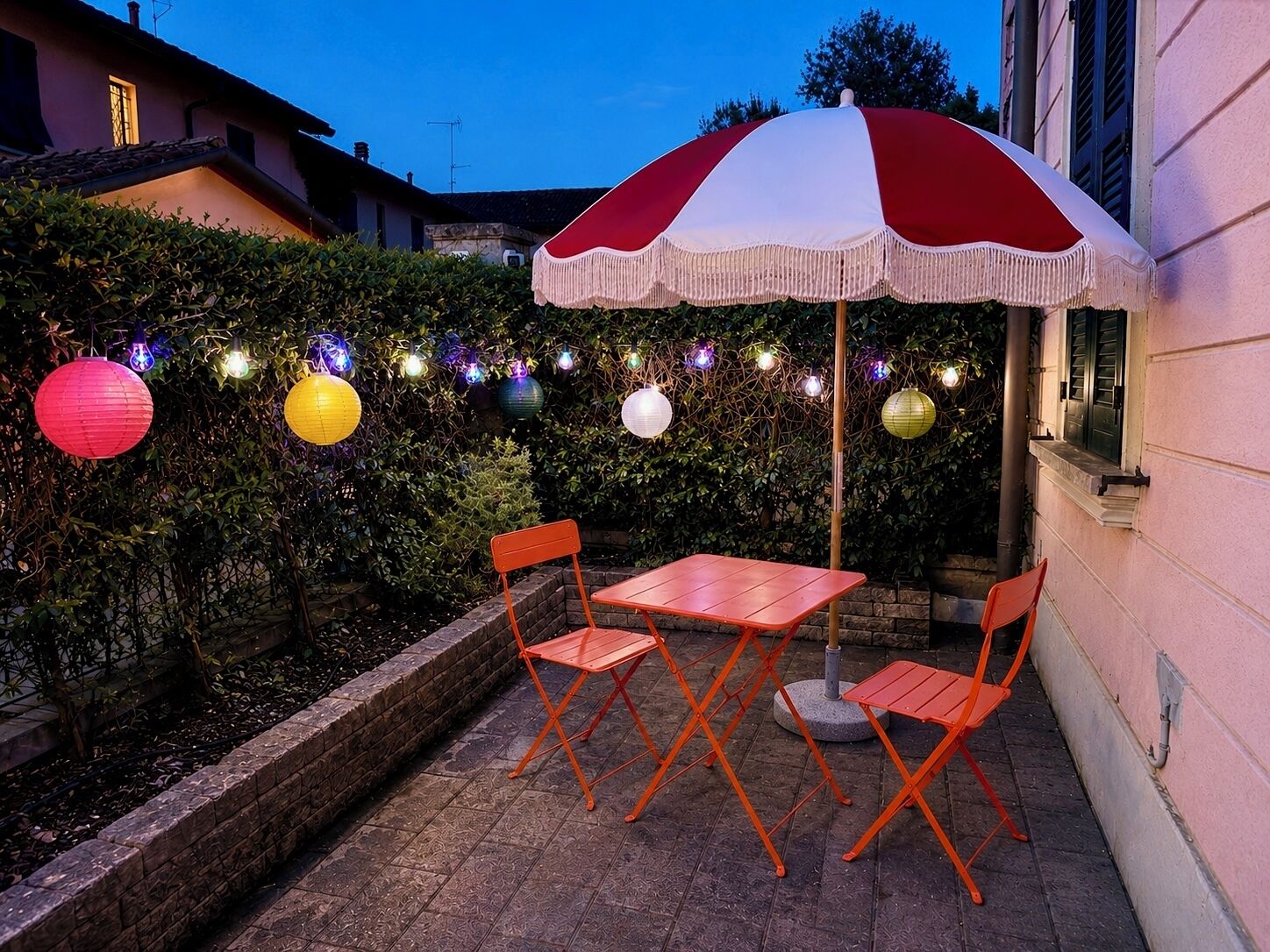 Un patio al aire libre con mesa, sillas, luces colgantes y un paraguas decorativo.