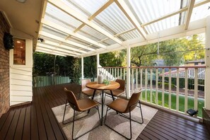 A covered patio with a wooden deck, a round table with chairs, and a view of a green garden. - Charming Family Home | Pool & Entertainer's Deck (Carlingford)
