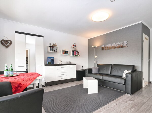 A modern living room with a grey sofa, a white coffee table, and a dining area with a red tablecloth. There are bookshelves with books and decorative items, and a large mirror on the wall. - Ankertied: Your cozy vacation apartment in Büsum, near the sea (Büsum)