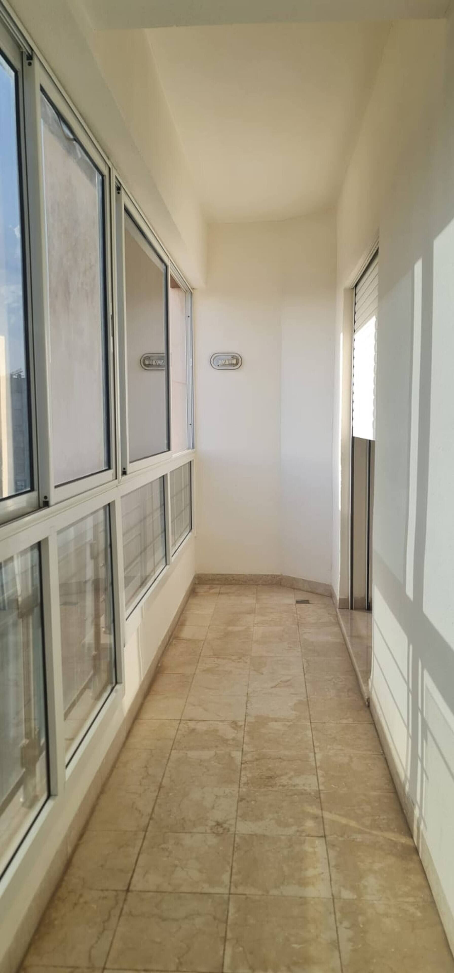 A narrow corridor with tiled flooring, large windows on one side, and a door on the other.