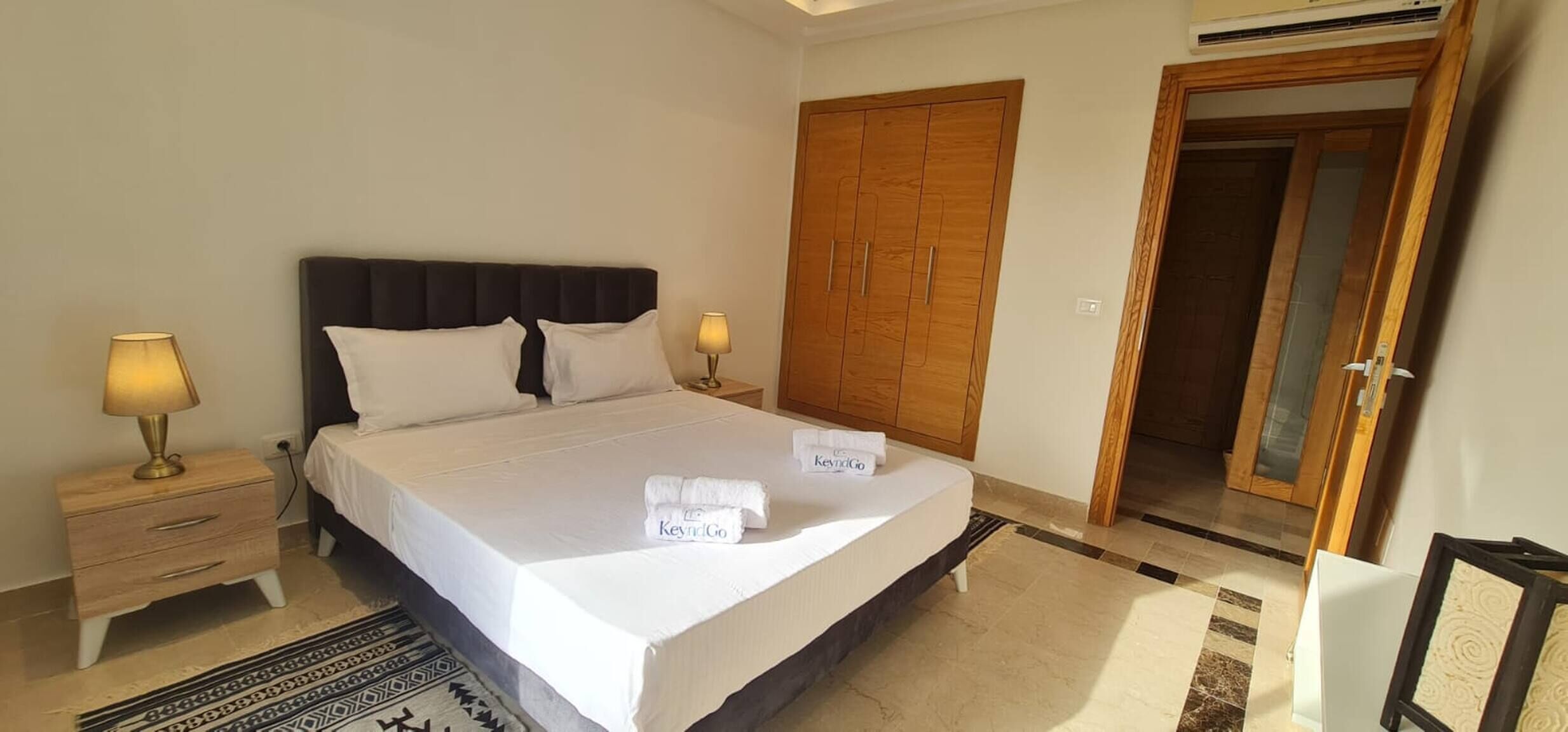 Habitación de hotel con cama, dos lámparas de noche, un armario de madera y una puerta que da a otra habitación.