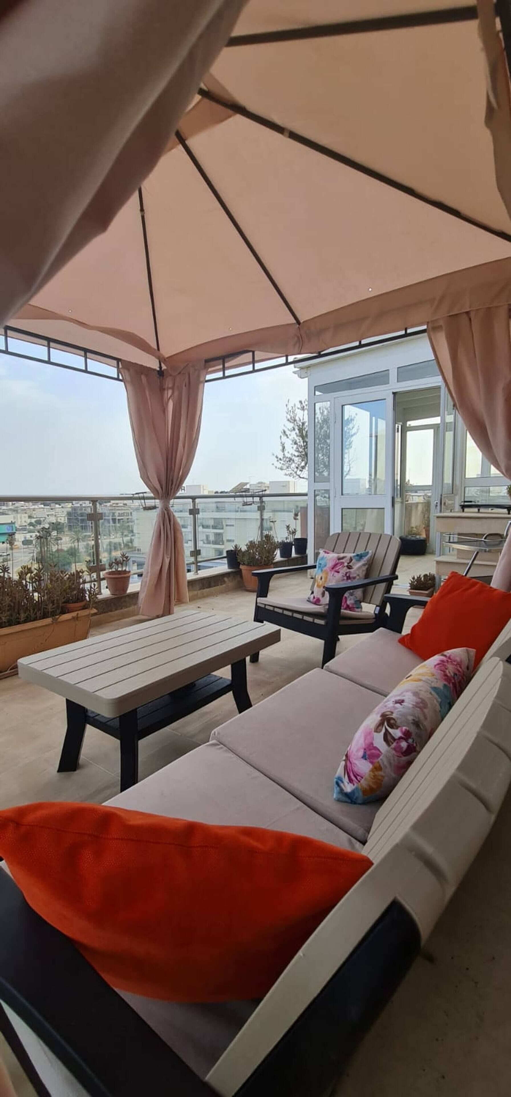A covered outdoor seating area with a table and lounge chairs, overlooking a cityscape.