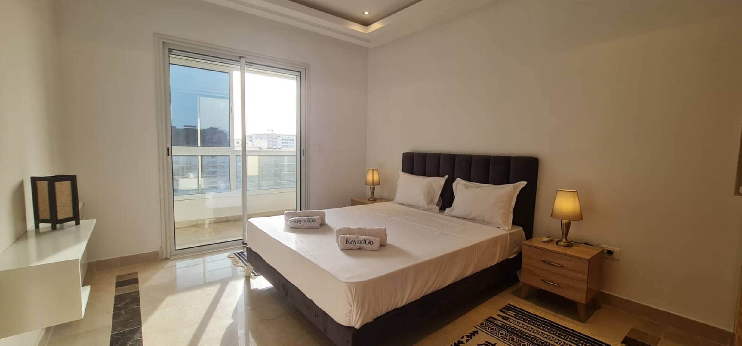 A hotel room with a large bed, two bedside lamps, a view through a sliding door, and a small table with a lamp.