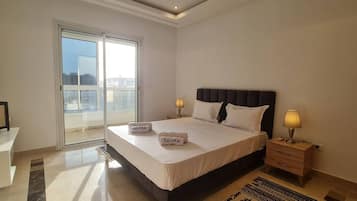 A hotel room with a large bed, two bedside lamps, a view through a sliding door, and a small table with a lamp.