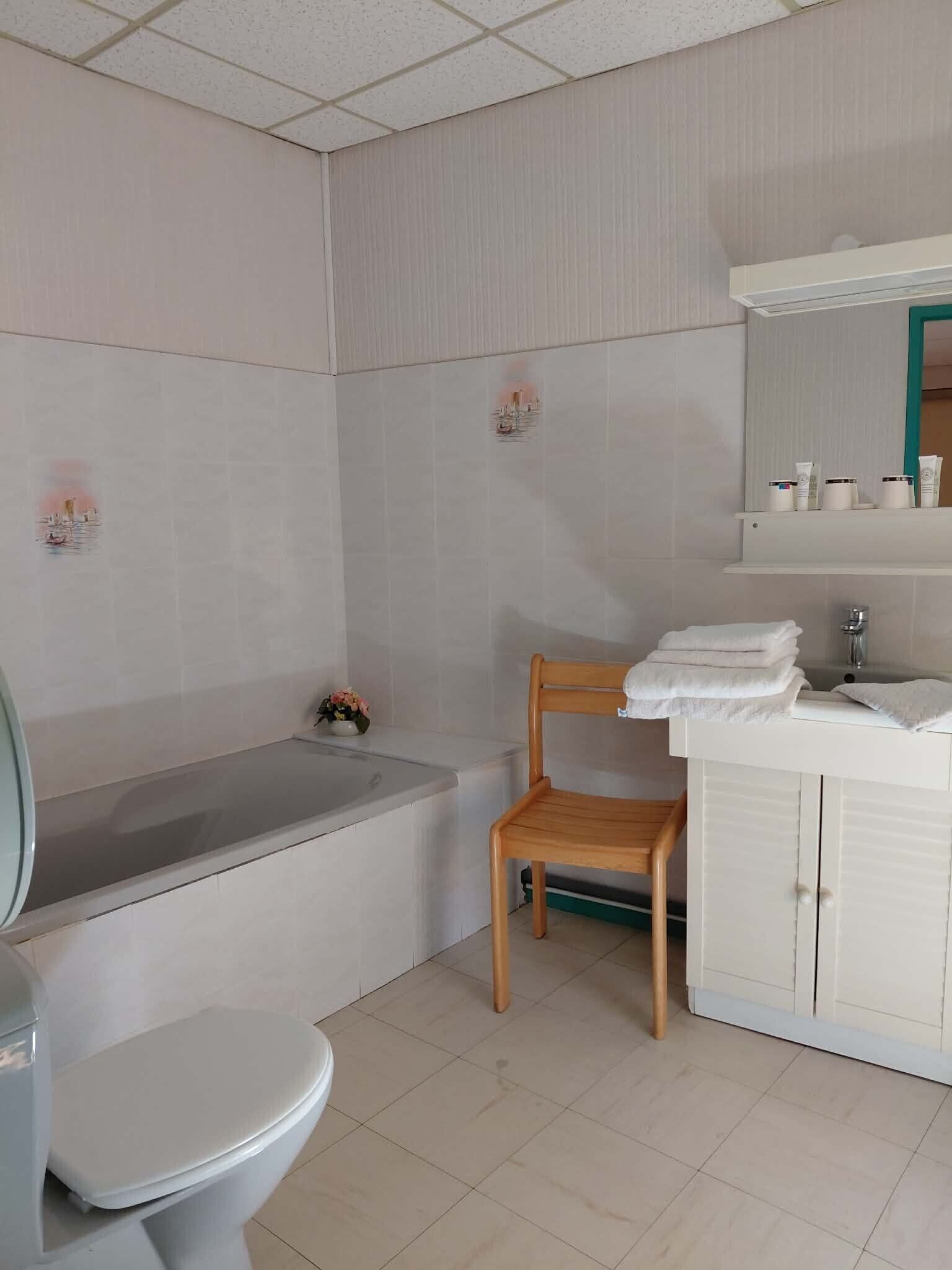 A bathroom with a toilet, bathtub, wooden chair, and a sink area with toiletries.