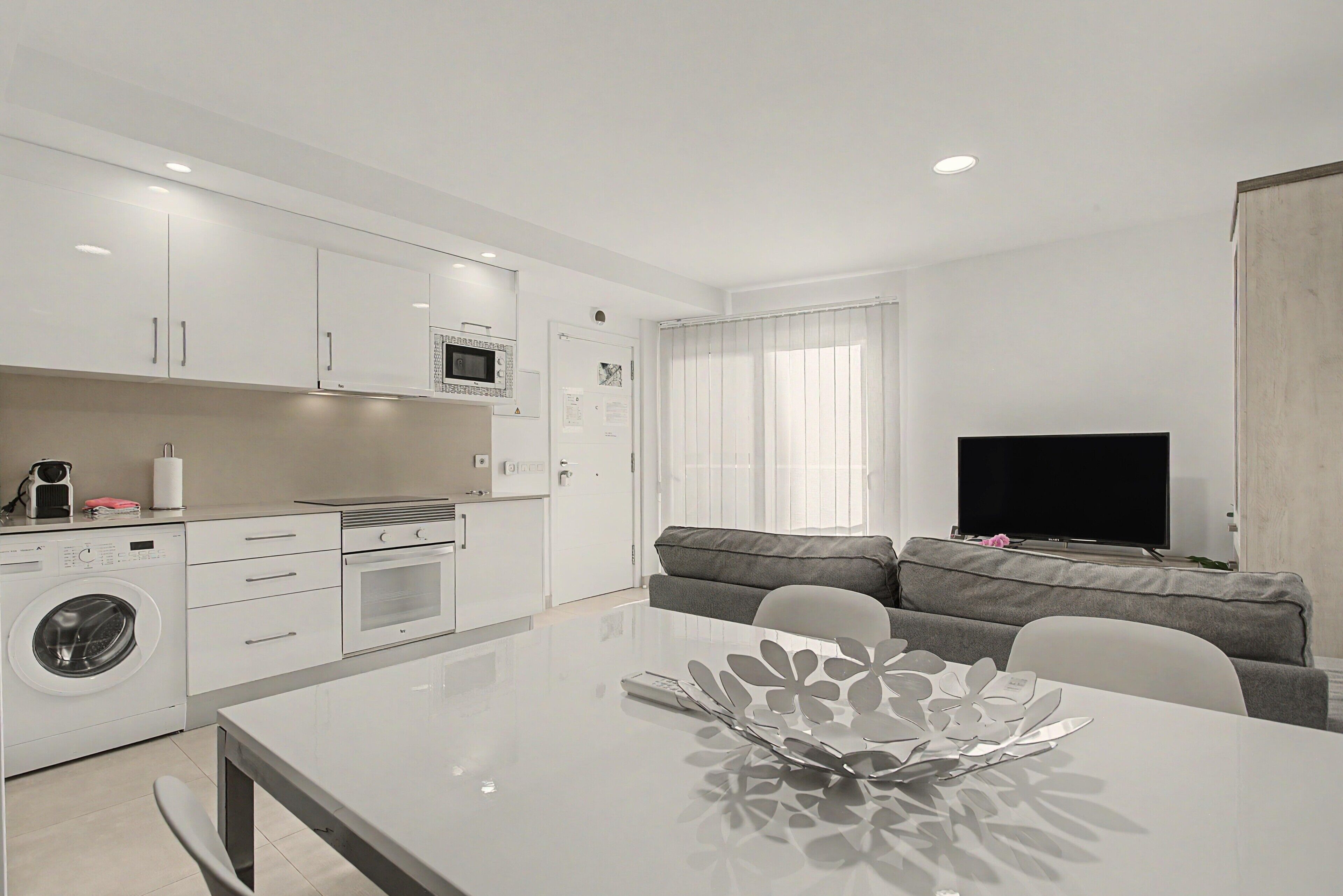 Een moderne keuken met een ingebouwde magnetron, oven en wasmachine. Er is een eettafel met een decoratief middelpunt. In de woonkamer staat een grijze bank en een flatscreen televisie.