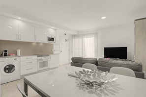 A modern kitchen with a built-in microwave, oven, and washing machine. There is a dining table with a decorative centerpiece. A grey sofa and a flat-screen TV are in the living area. - Vacation Apartment 'Brisa C' with Balcony, Wi-Fi and Air Conditioning (El Médano)