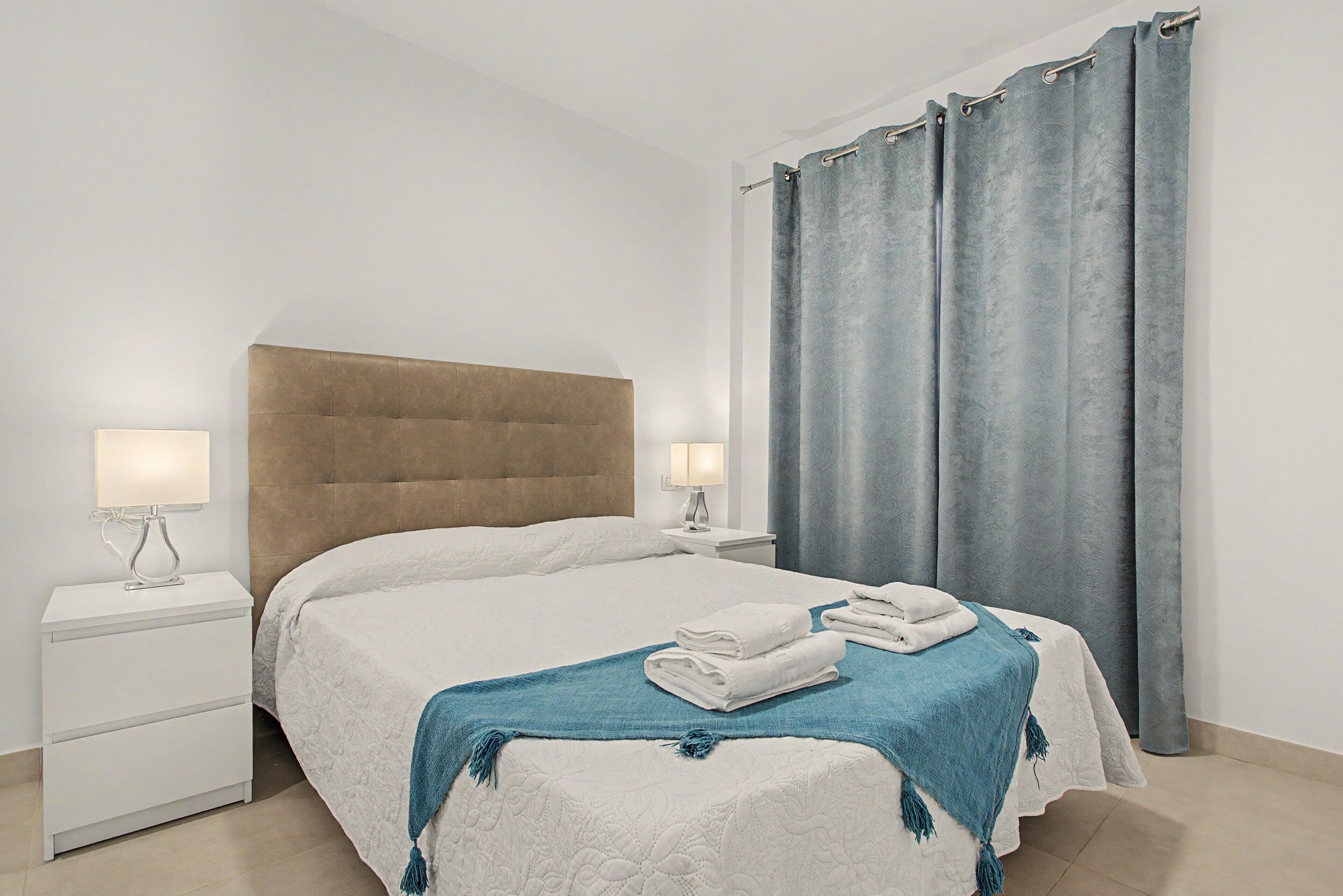 Una habitación de hotel con una cama, dos mesitas de noche, una lámpara y un ventanal con cortinas.