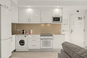 Cocina compacta equipada con lavadora, horno, microondas y una pequeña zona de comedor con sofá.
