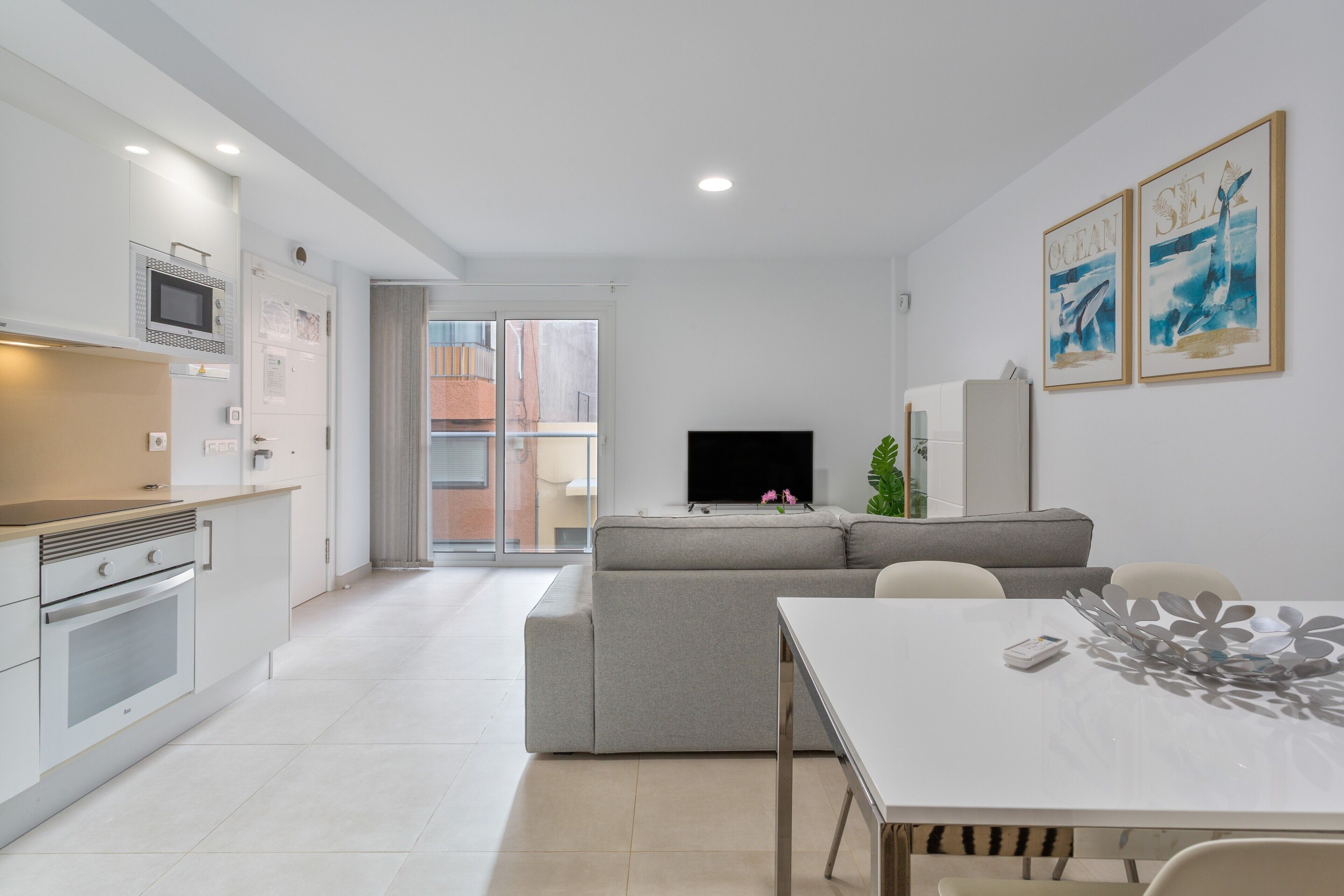 Una cocina moderna con horno empotrado, un comedor con mesa y sillas, un sofá gris y un televisor de pantalla plana montado en la pared.