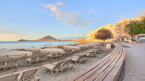 A beach with rows of sun loungers and umbrellas, a clear sky, and a mountain in the distance. - Vacation Apartment 'Brisa A. Apto En El Médano' with Wi-Fi and Air Conditioning (El Médano)