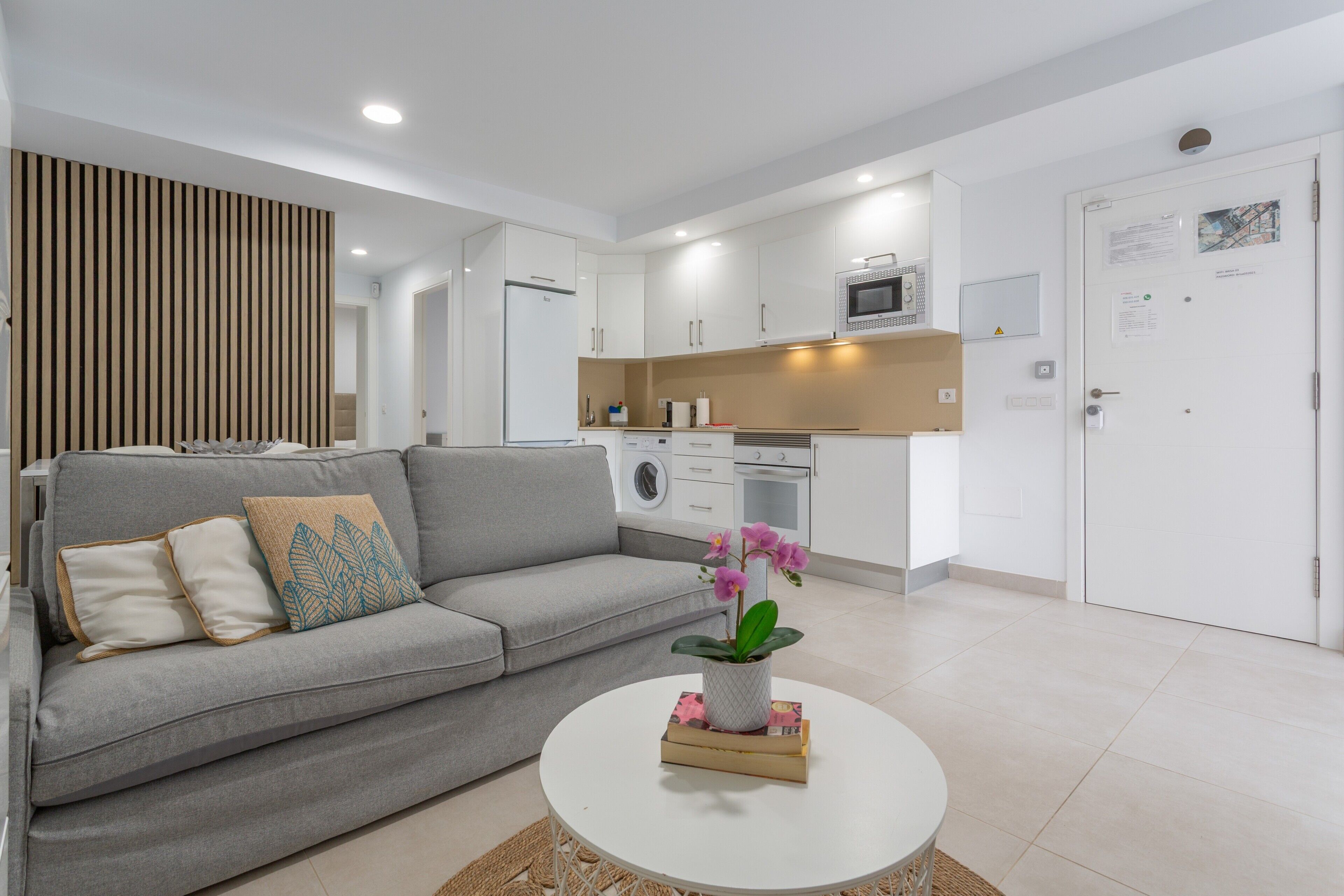 Una sala de estar moderna con un sofá gris, una mesita de centro con una planta y una cocina al fondo.