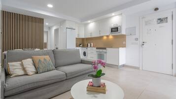 Una sala de estar moderna con un sofá gris, una mesita de centro con una planta y una cocina al fondo.