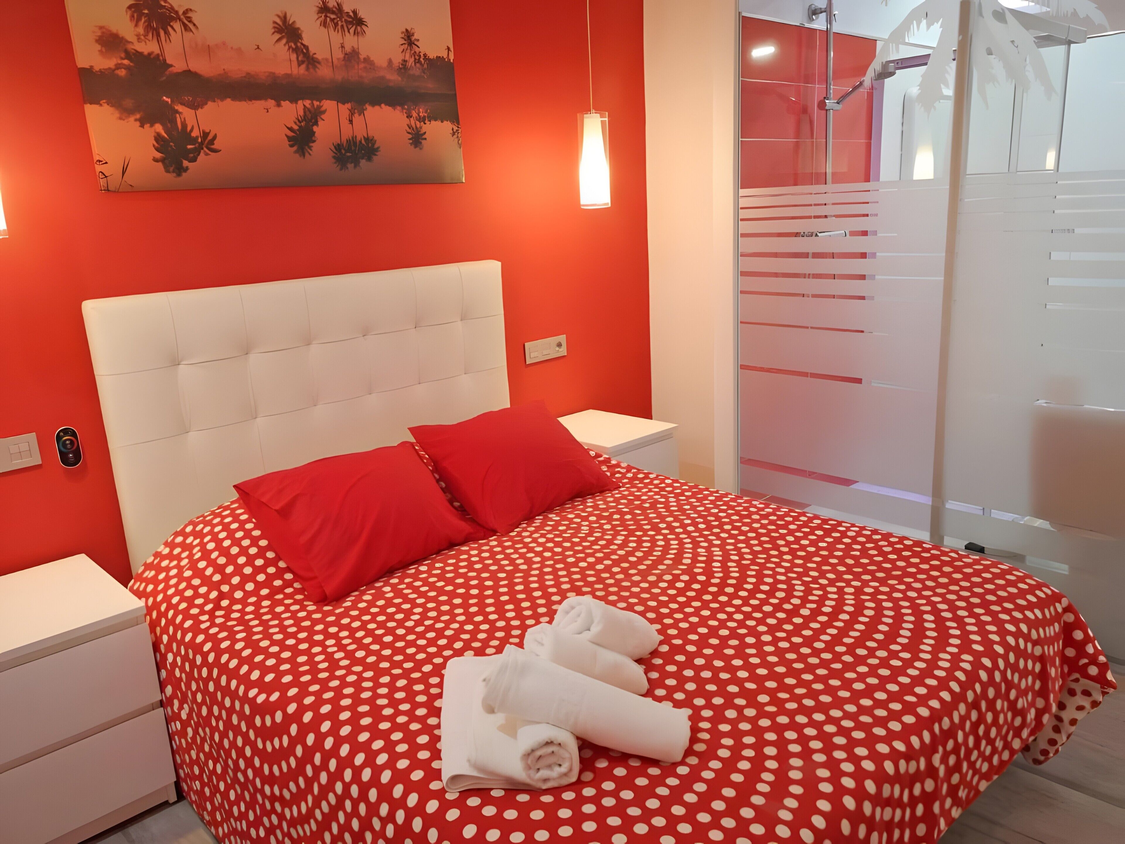 Habitación de hotel con colcha roja de lunares, cabecera blanca, almohadas rojas y un cuadro de palmeras en la pared.
