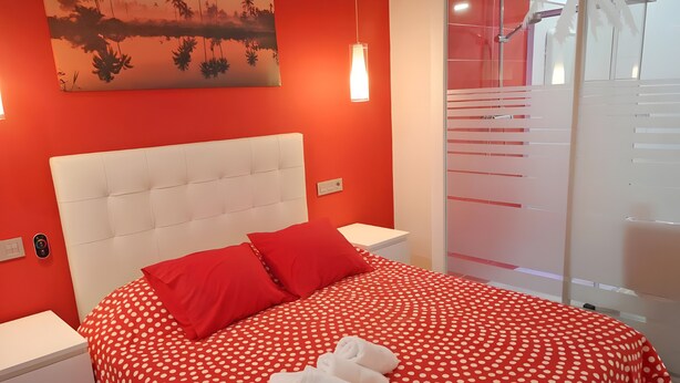 A hotel room with a red polka dot bedspread, white headboard, red pillows, and a painting of palm trees on the wall. - Studio Apartment 'Ofis - Estudio Rojo' with Wi-Fi and Air Conditioning (Conil de la Frontera)