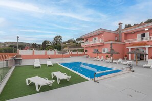 Un bâtiment à deux étages doté d’une piscine, de chaises longues blanches et d’un parasol en chaume.