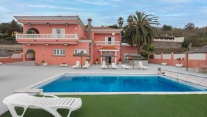 A two-story building with a swimming pool, white lounge chairs, and a palm tree. - 4-Bedroom Villa El Romero—Private Pool, BBQ & Relaxation (Arucas)