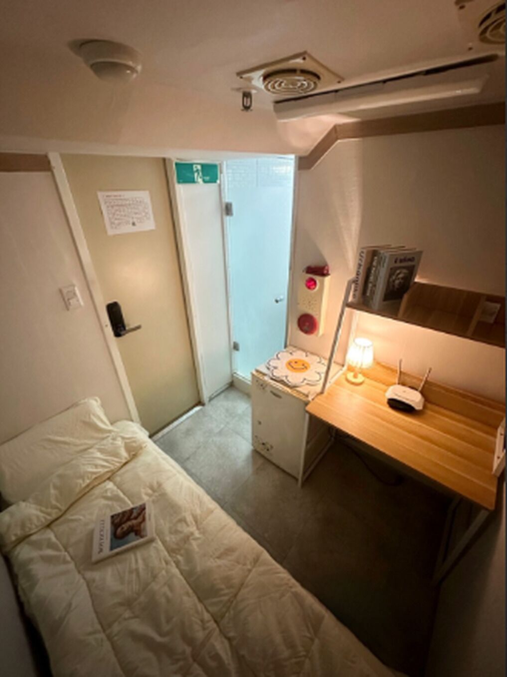 A small, compact room with a single bed, a desk with a lamp, and a small table with a device. There is a door with a sign on it, and a small cabinet or storage unit.
