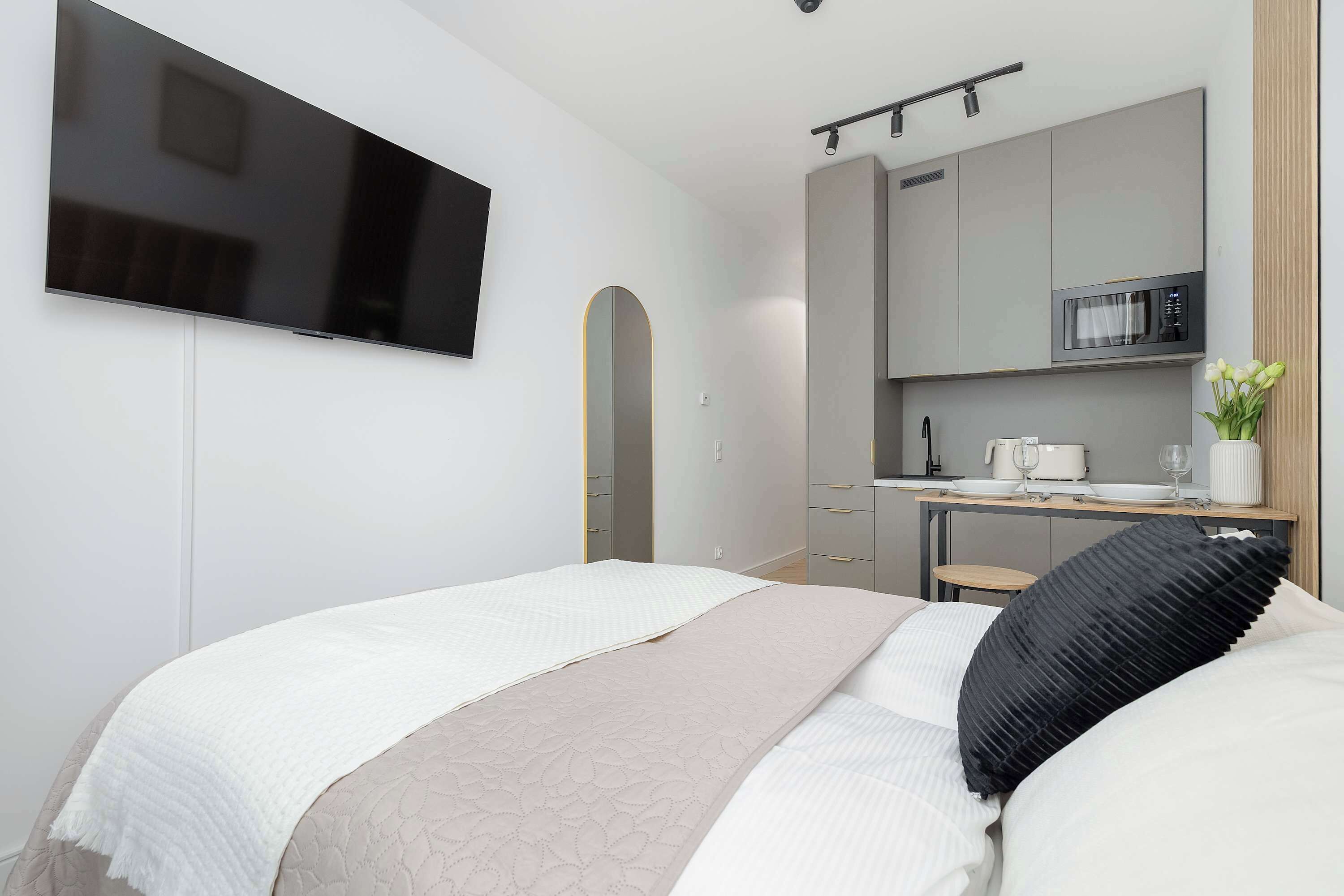 Moderni hotellihuone, jossa on sänky, taulutelevisio, pieni keittiönurkkaus ja pieni pöytä, jonka päällä on maljakko täynnä kukkia.