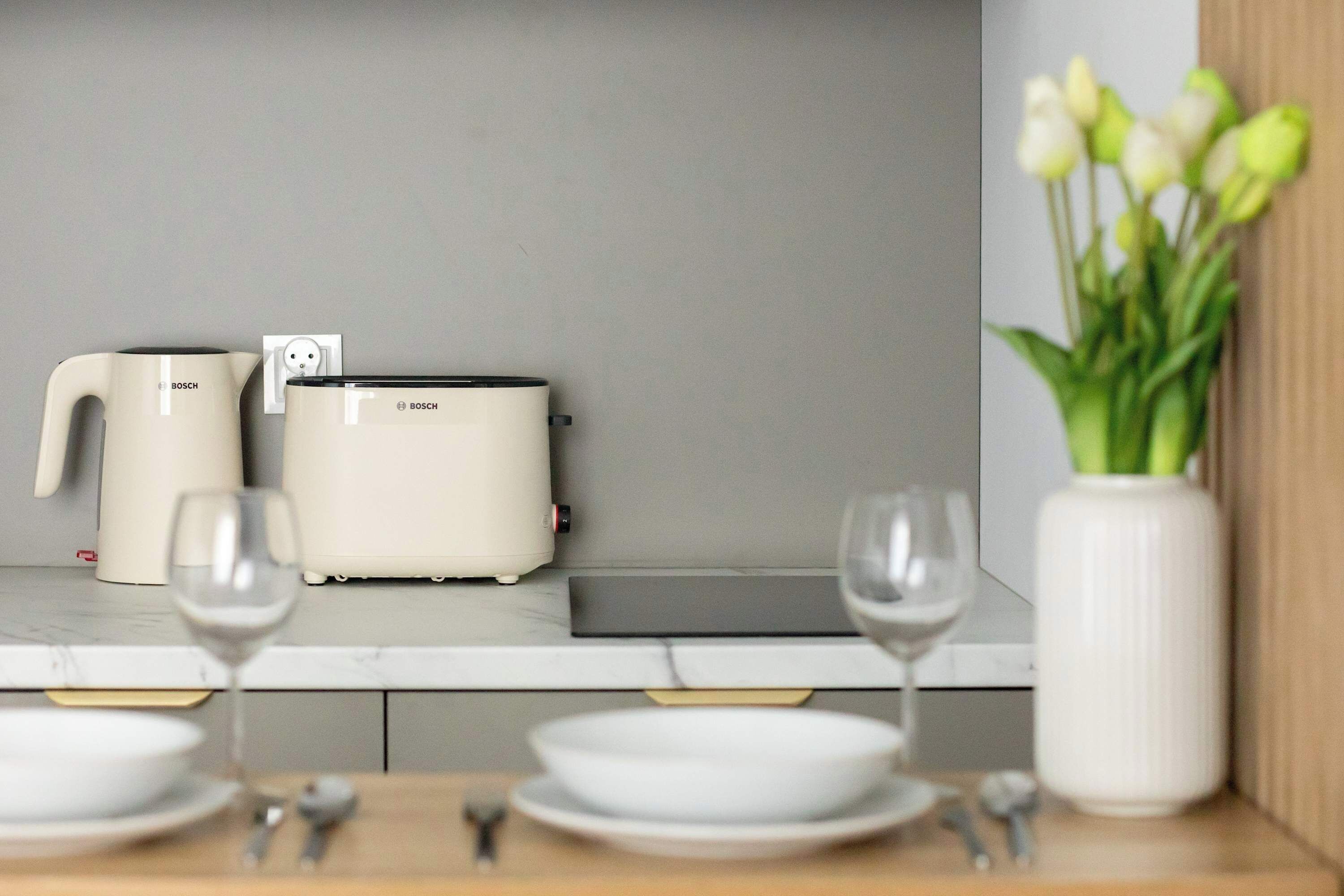 Uma cozinha com uma torradeira e chaleira Bosch, um vaso com flores e uma mesa de jantar posta com pratos e copos.