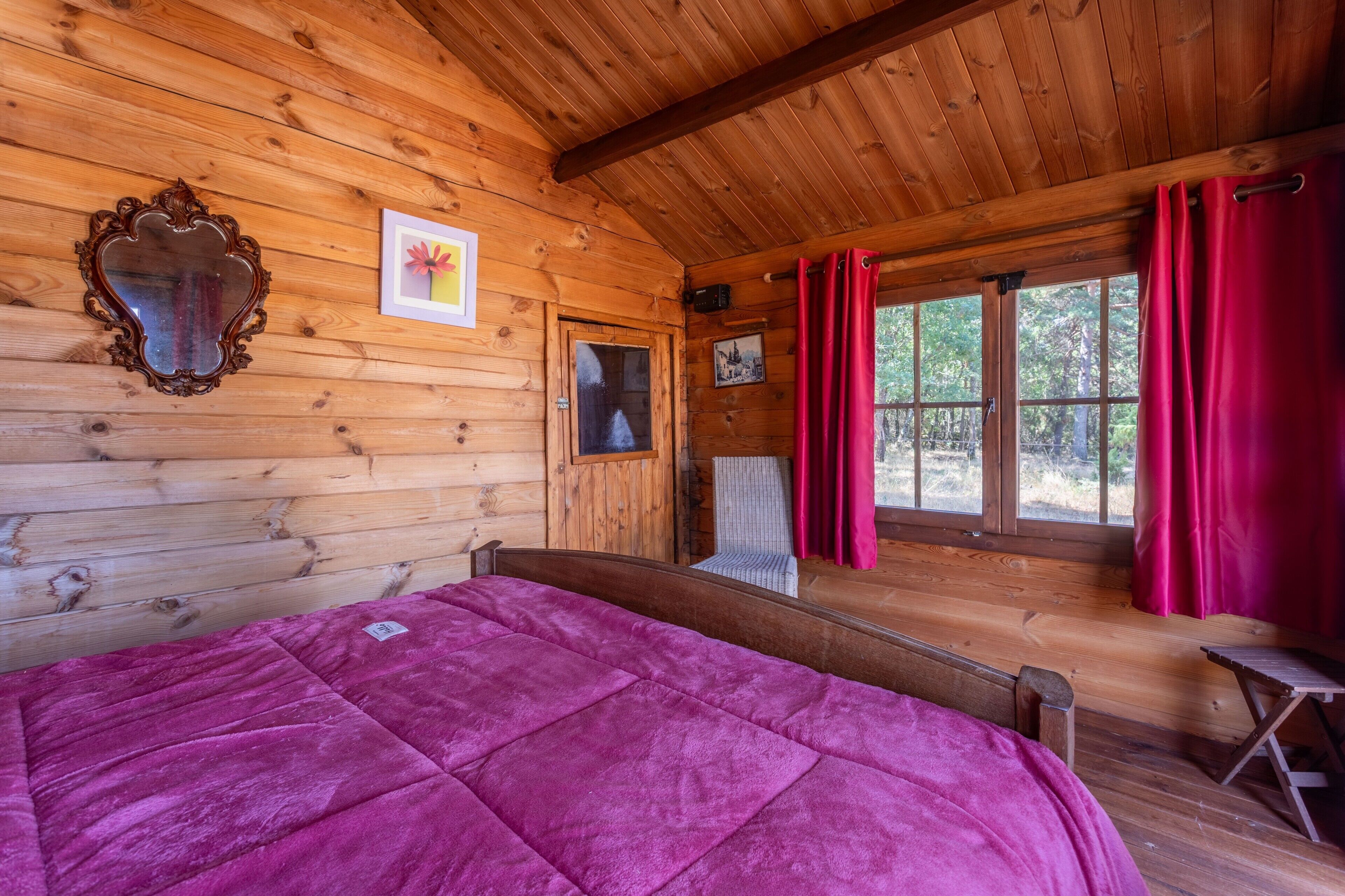 A wooden cabin room with a bed, a chair, a window with curtains, and a mirror on the wall.