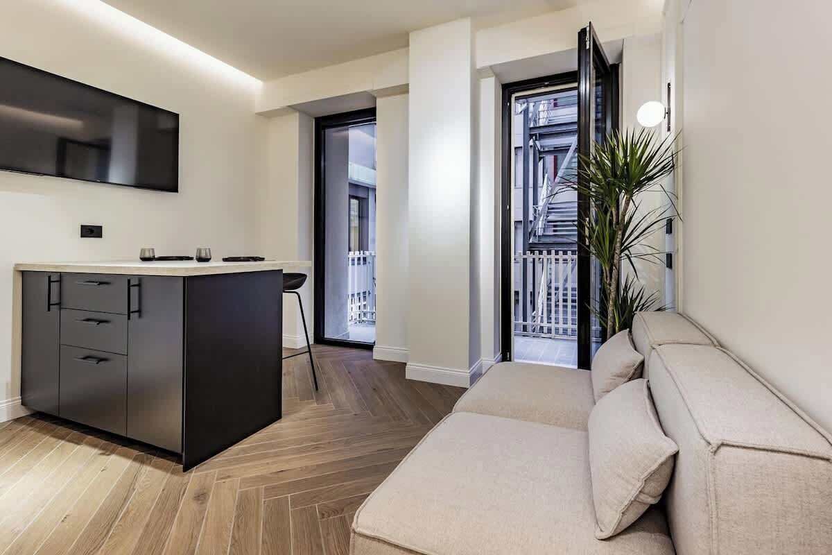 A modern kitchen with a black countertop and cabinets, a large flat-screen TV mounted above the counter, and a wooden herringbone floor.