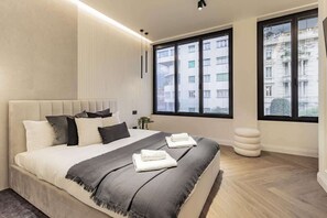 A modern bedroom with a large bed, a bedside table, a window with blinds, and a view of buildings outside. - [Lungolago] Nuovo Bilocale 5 (Como)