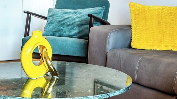 A glass-top coffee table with a yellow object on it, a teal armchair with a cushion, and a brown sofa with a yellow pillow. - silk and sunset - city and lake view (Como)