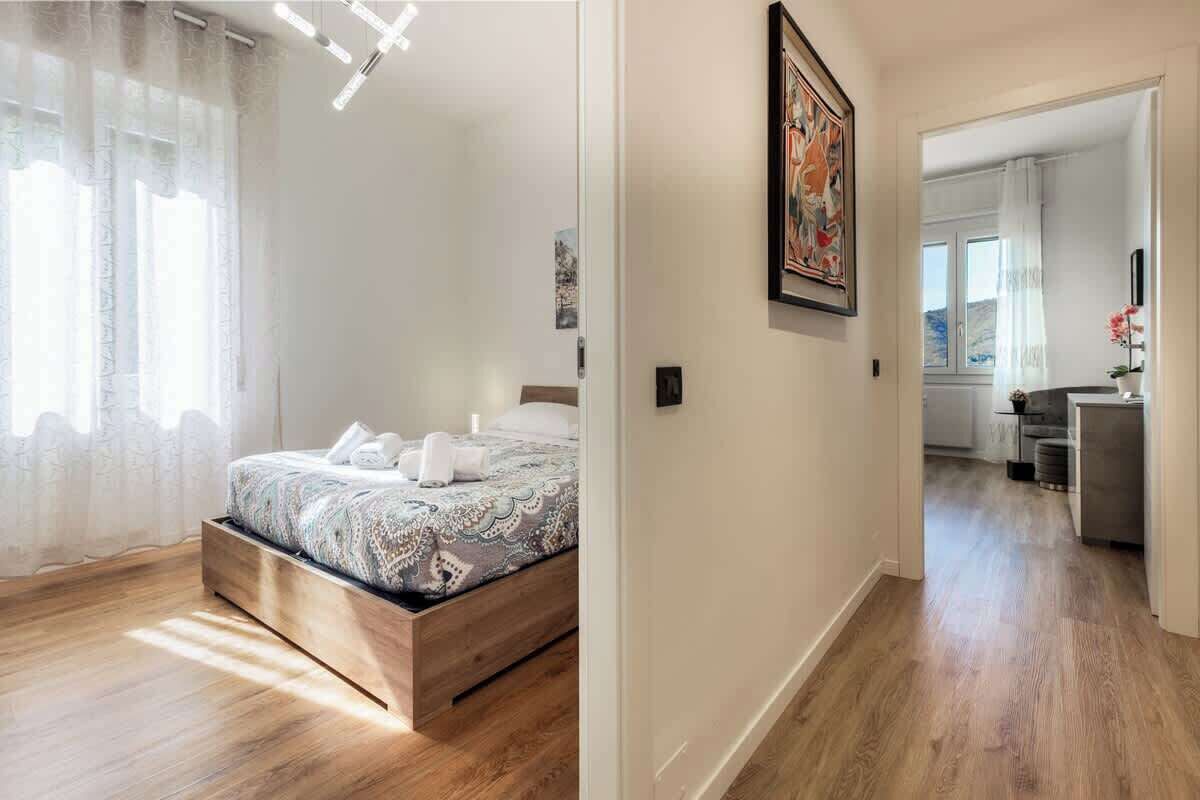 Una camera da letto con un letto in legno, un'ampia finestra con tende leggere e un corridoio che conduce al bagno.